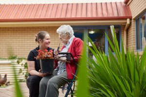 Female home care worker holding a pot plant and kneeling next to female Home Care Package client sitting on a walker